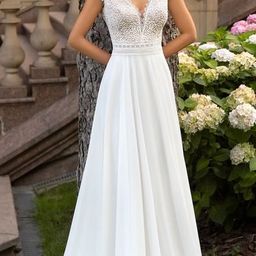 Eine lächelnde Frau in einem weißen Hochzeitskleid mit Spitzendetails steht neben einem Baum in der Nähe einer Treppe mit blühenden Blumen im Hintergrund.