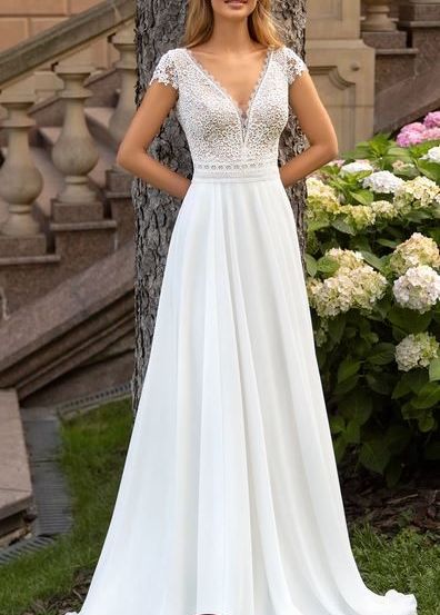 Eine lächelnde Frau in einem weißen Hochzeitskleid mit Spitzendetails steht neben einem Baum in der Nähe einer Treppe mit blühenden Blumen im Hintergrund.