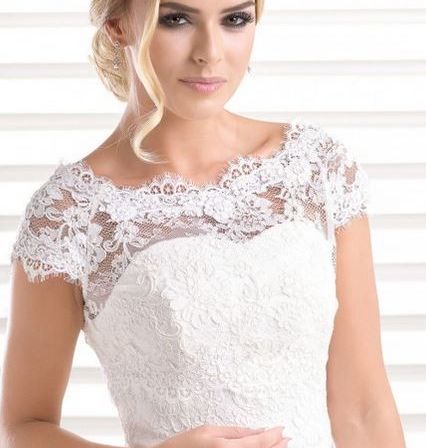 Eine Frau in einem weißen Hochzeitskleid mit Spitzendetails posiert für ein Foto und lächelt mit den Händen auf der Taille.