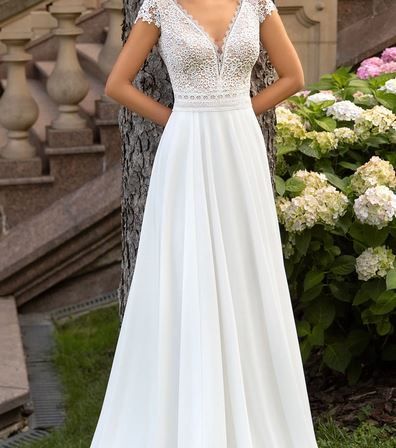 Eine Frau steht auf Gras neben einem Baum in einem weißen, spitzenbesetzten Hochzeitskleid. Sie posiert lächelnd. Hinter ihr befindet sich eine Treppe mit Geländern und verschiedenen Blumen.