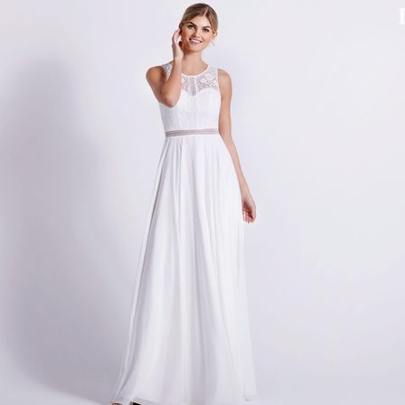 Eine Frau trägt ein weißes, ärmelloses Kleid mit Spitzendetails am Ausschnitt und Taille, sie steht vor einer weißen Wand.