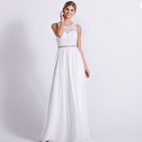 Eine Frau trägt ein weißes, ärmelloses Kleid mit Spitzendetails am Ausschnitt und Taille, sie steht vor einer weißen Wand.