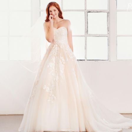 Eine Frau in einem weißen Hochzeitskleid steht lächelnd vor einem Fenster. Sie hat einen Schleier auf dem Kopf.