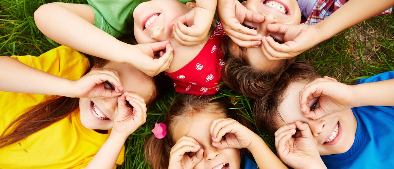 Mehrere kleine Kinder liegen auf einem Grasfeld und bilden einen Kreis. Sie alle lächeln und halten ihre Hände vor die Augen.