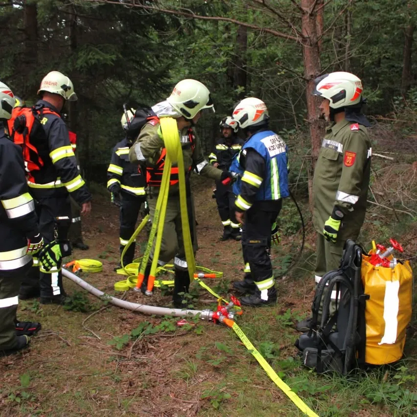 Eine Gruppe Feuerwehrleute in einem Wald steht herum, spricht und hält Werkzeuge. Sie tragen Helme und Schutzanzüge. Ein gelber Beutel und Schläuche liegen auf dem Boden.