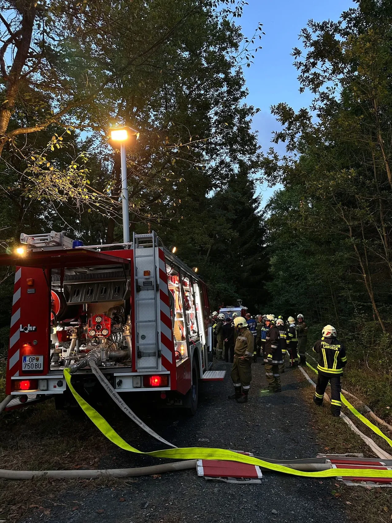 Ein Feuerwehrauto ist am Straßenrand geparkt, Feuerwehrleute kümmern sich um einen Notfall. Das Feuerwehrauto hat eine offene Tür und eine angeschlossene Feuerwehrschläuche.
