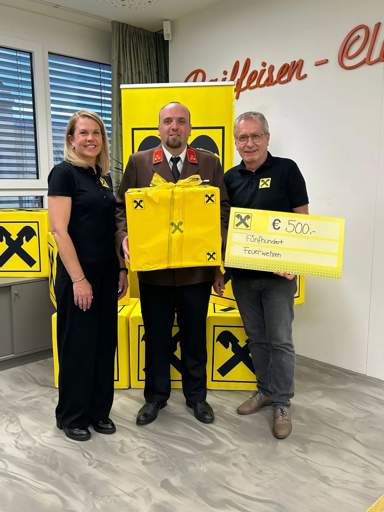 Drei Personen stehen in einem Raum mit gelben Boxen dahinter. Ein Mann in Uniform hält eine Geschenkbox und ein anderer Mann hält ein Zertifikat mit 500 Euro. Eine Frau steht neben ihnen.