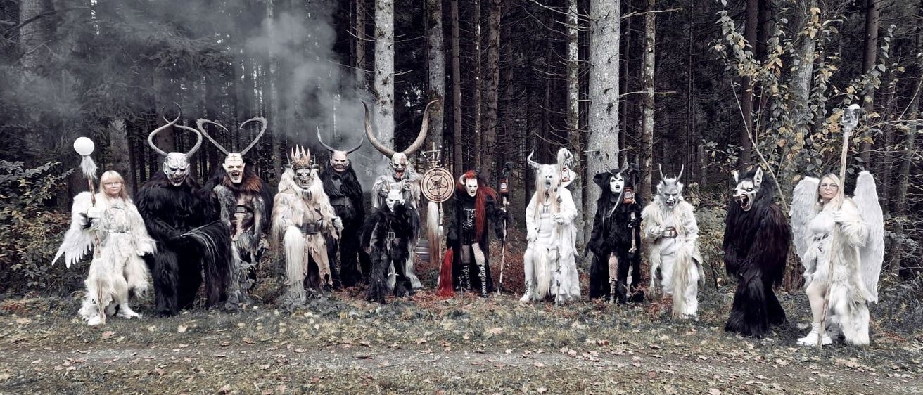 Eine Gruppe von Personen in aufwendigen Kostümen, einschließlich Masken und Hörnern, steht in einer nebligen Waldumgebung.