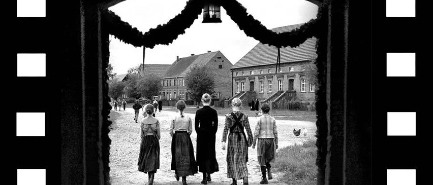 Ein Schwarz-Weiß-Foto von fünf Kindern, die auf einer Straße vor Gebäuden gehen, eines hält ein Huhn, und mehrere Personen gehen im Hintergrund.