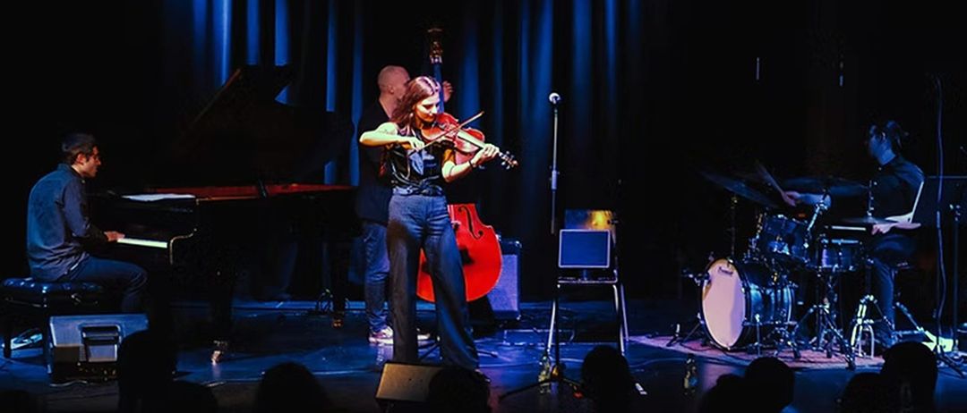 Eine Frau spielt Geige auf der Bühne, begleitet von einem Mann, der Cello spielt. Sie stehen vor einem Klavier, einem Mikrofonständer und einem Laptop. Vor ihnen sitzt ein Publikum.