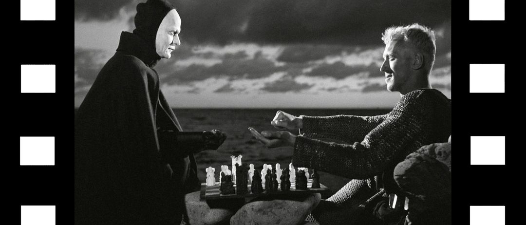 Zwei Männer spielen Schach an einem felsigen Ufer unter einem bewölkten Himmel. Einer trägt eine Kapuze und der andere ist in Rüstung.