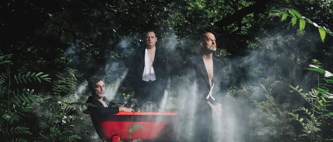 Drei Männer befinden sich in einem Wald. Einer sitzt in einer roten Wanne, während zwei weitere hinter ihm stehen, alle in Schwarz gekleidet. Rauch umgibt sie.