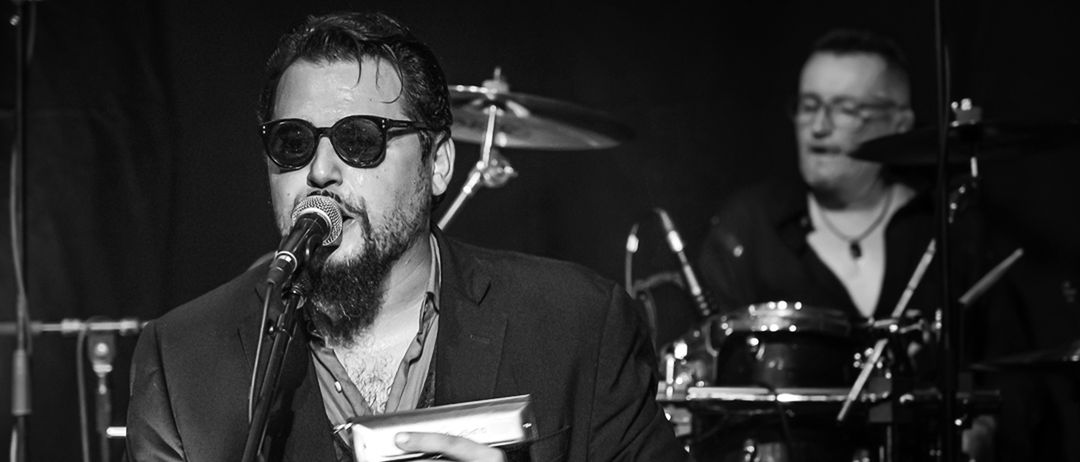 Ein bärtiger Mann mit Sonnenbrille singt in ein Mikrofon auf der Bühne. Dahinter steht ein Schlagzeug mit Becken.