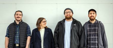 Ein Mann und eine Frau stehen Seite an Seite, lächelnd, vor einer schlichten weißen Wand. Der Mann trägt eine schwarze Jacke mit weißem Innenshirt und eine Brille, während die Frau einen marineblauen Mantel und eine Brille trägt.