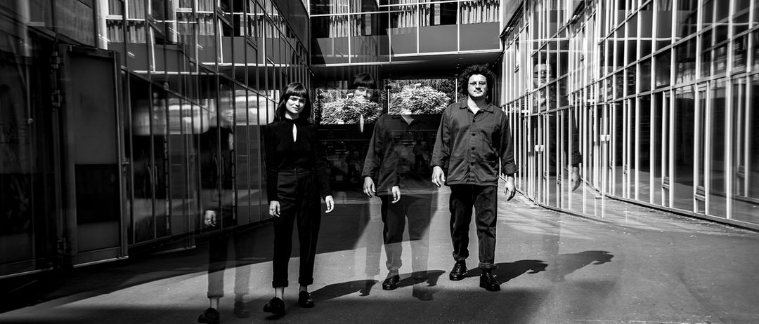 Drei Personen stehen in einem Schwarz-Weiß-Foto vor einem Gebäude mit Glaswänden und Vorhängen. Die Person auf der rechten Seite trägt eine Brille.