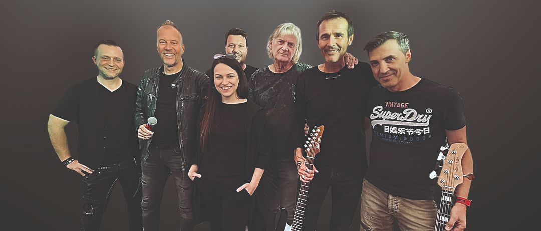 Eine Gruppe von Musikern, bestehend aus fünf Männern und einer Frau, lächeln und posieren für ein Foto. Sie stehen eng beieinander.