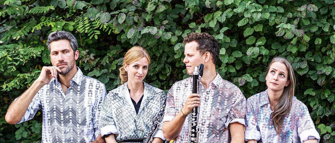Drei Musiker stehen vor einem grünen Busch. Die Frau in der Mitte lächelt, während der Mann rechts eine Klarinette hält.