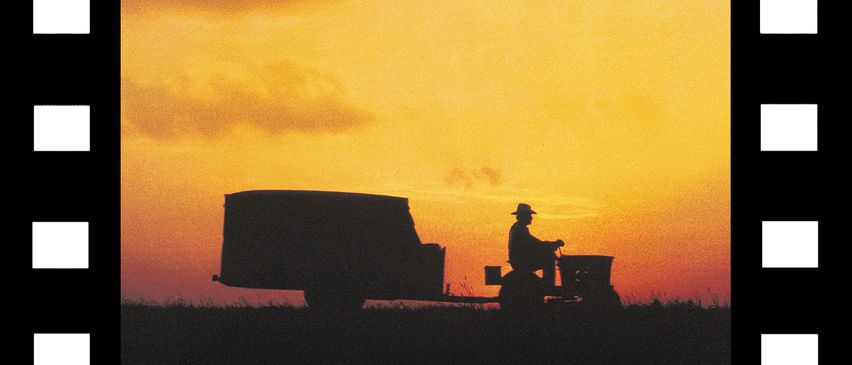 Eine Silhouette eines Bauern, der einen Traktor mit Anhänger fährt, bei Sonnenuntergang mit einem warmen Himmel und Wolken.