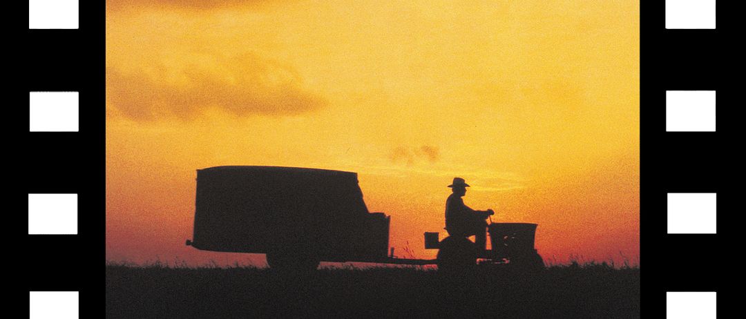 Eine Silhouette eines Bauern, der einen Traktor mit Anhänger fährt, bei Sonnenuntergang mit einem warmen Himmel und Wolken.