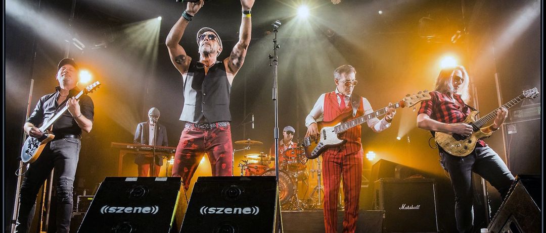 Eine Band tritt auf der Bühne auf, wobei der Leadsänger von einem Scheinwerfer beleuchtet wird, der ein Mikrofon hält. Hinter ihm spielt ein Gitarrist eine E-Gitarre. Ein Schlagzeuger und ein Keyboarder sind im Hintergrund. Die Bühne ist mit Lautsprechern und Bühnenbeleuchtung ausgestattet.