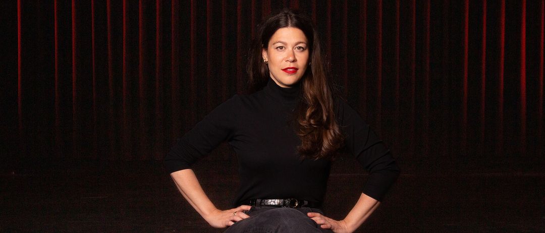 Eine Frau mit langen, lockigen Haaren sitzt auf einem Sofa und trägt ein schwarzes Rollkragenshirt und Jeans. Sie hat die Hände in die Hüften gestemmt und schaut direkt in die Kamera. Sie hat roten Lippenstift auf.