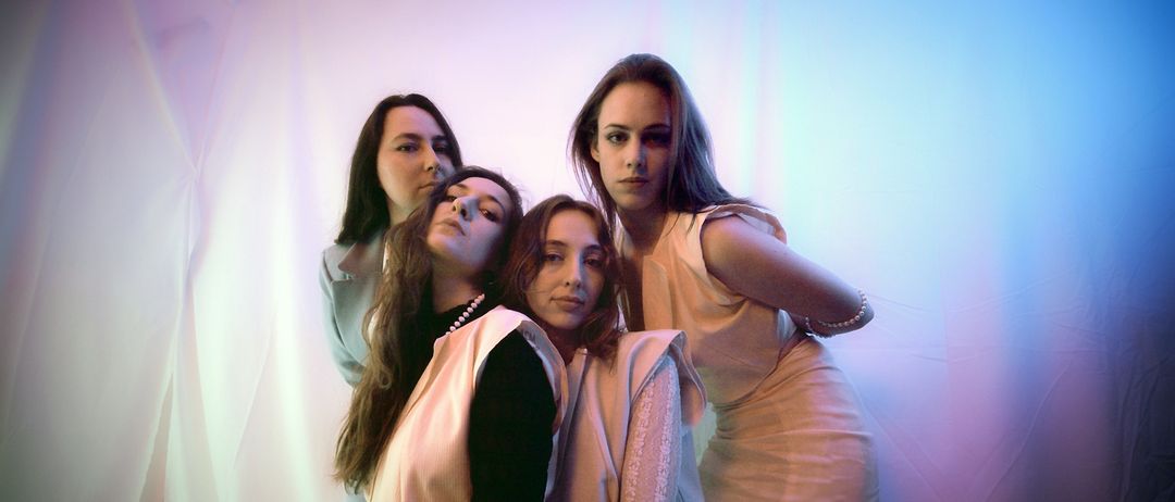 Vier Frauen posieren zusammen in einem Studio, eine hat einen ernsten Gesichtsausdruck, während die anderen verspielte Gesichter machen. Sie tragen passende pastellfarbene Kleider und eine hat eine Perlenkette.