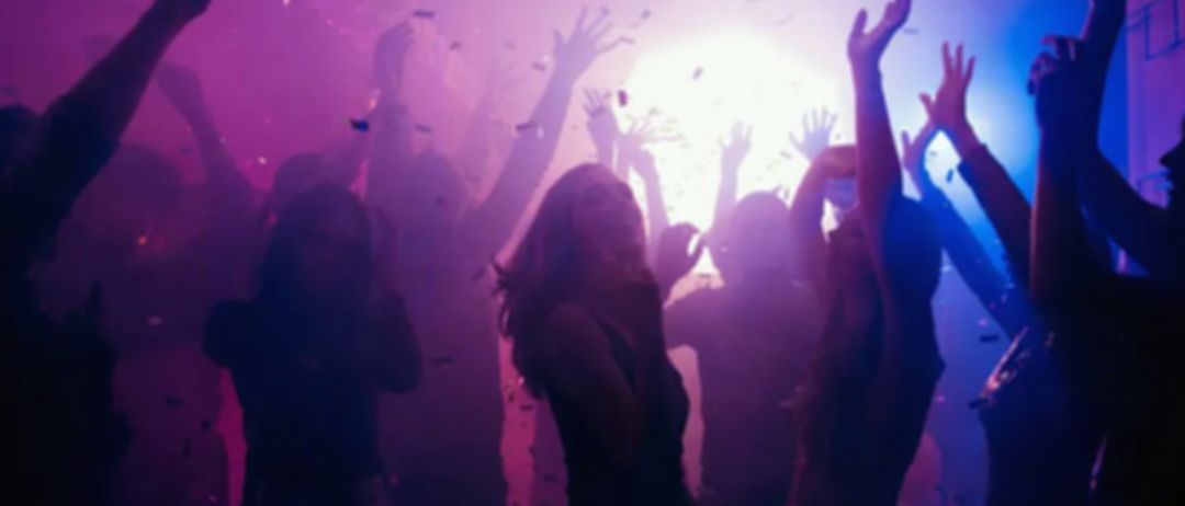 Eine Menschenmenge tanzt auf einer Party mit lila und rosa Lichtern und fallendem Konfetti.