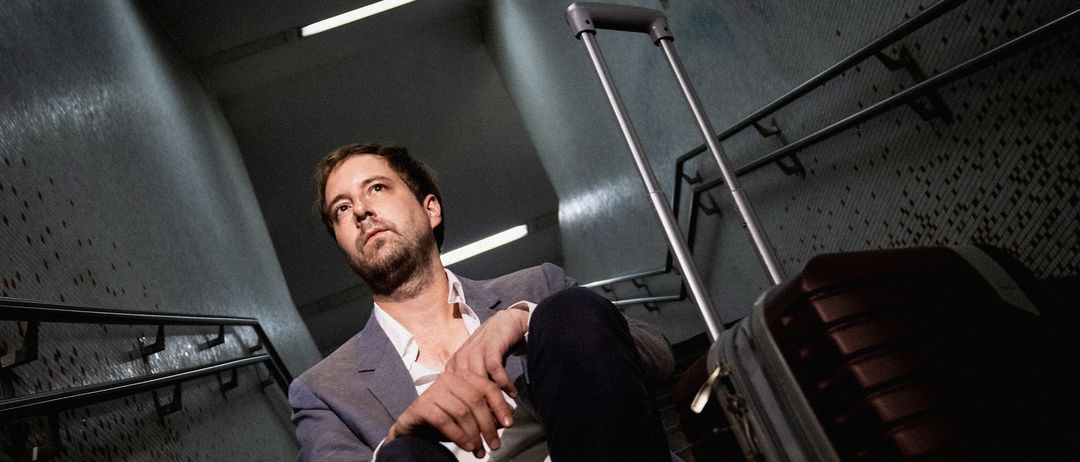 Ein Mann im Anzug sitzt auf den Stufen und schaut nachdenklich zur Seite. Neben ihm steht ein Koffer mit Griff. Die Umgebung ist schwach beleuchtet mit Metallgeländern.