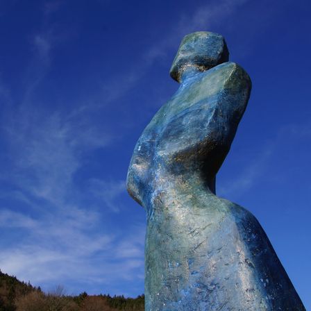 Eine große blaue Statue einer Frau steht vor einem klaren blauen Himmel, mit Bäumen und einem fernen Berg im Hintergrund.