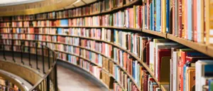 Ein Bibliotheksgang mit vielen Büchern auf Holzregalen, beleuchtet durch sanftes Licht von der Decke. Die Bücher sind in verschiedenen Farben und Größen angeordnet.