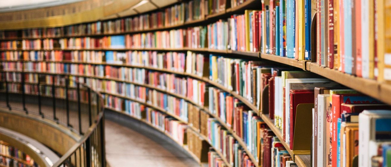 Ein Bibliotheksgang mit vielen Büchern auf Holzregalen, beleuchtet durch sanftes Licht von der Decke. Die Bücher sind in verschiedenen Farben und Größen angeordnet.
