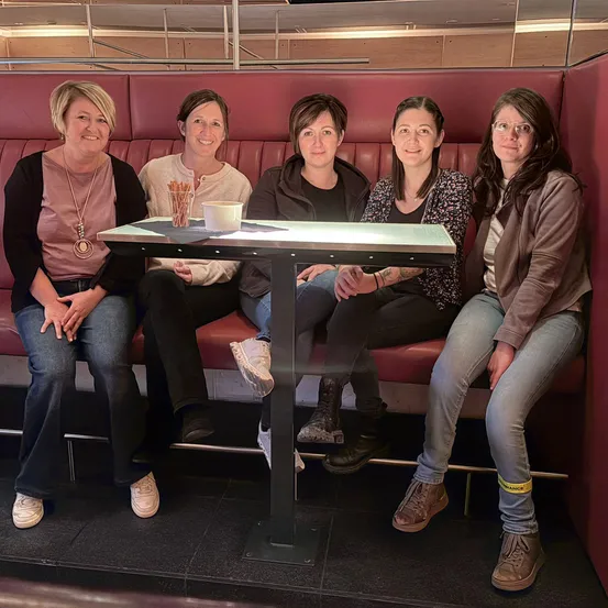 Fünf Frauen sitzen auf einer roten Bank in einem Café, lächeln und posieren für ein Foto. Sie tragen Freizeitkleidung. Vor ihnen steht ein kleiner Tisch mit einer Tasse und einem Untertasse.