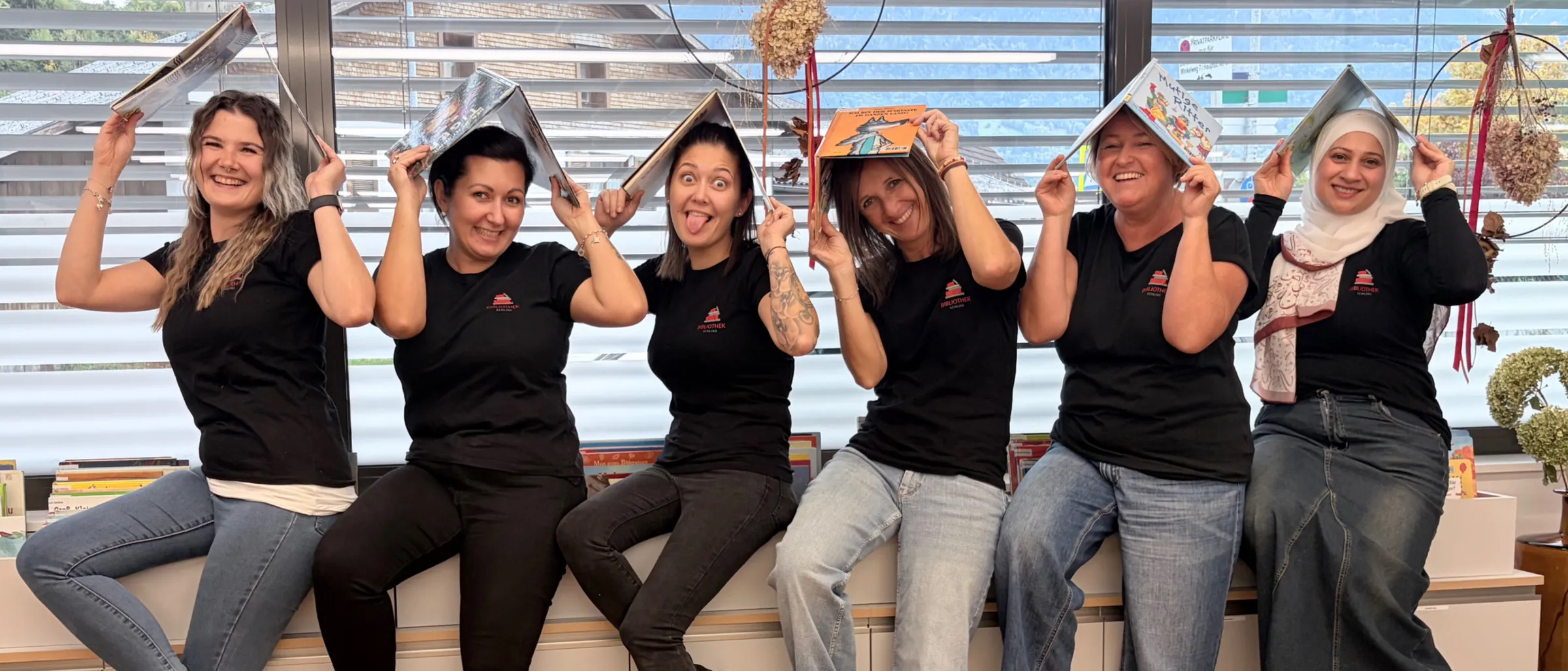 Eine Gruppe von Frauen in schwarzen T-Shirts und Jeans sitzt auf einer Bank, hält Bücher hoch und posiert für ein Foto.