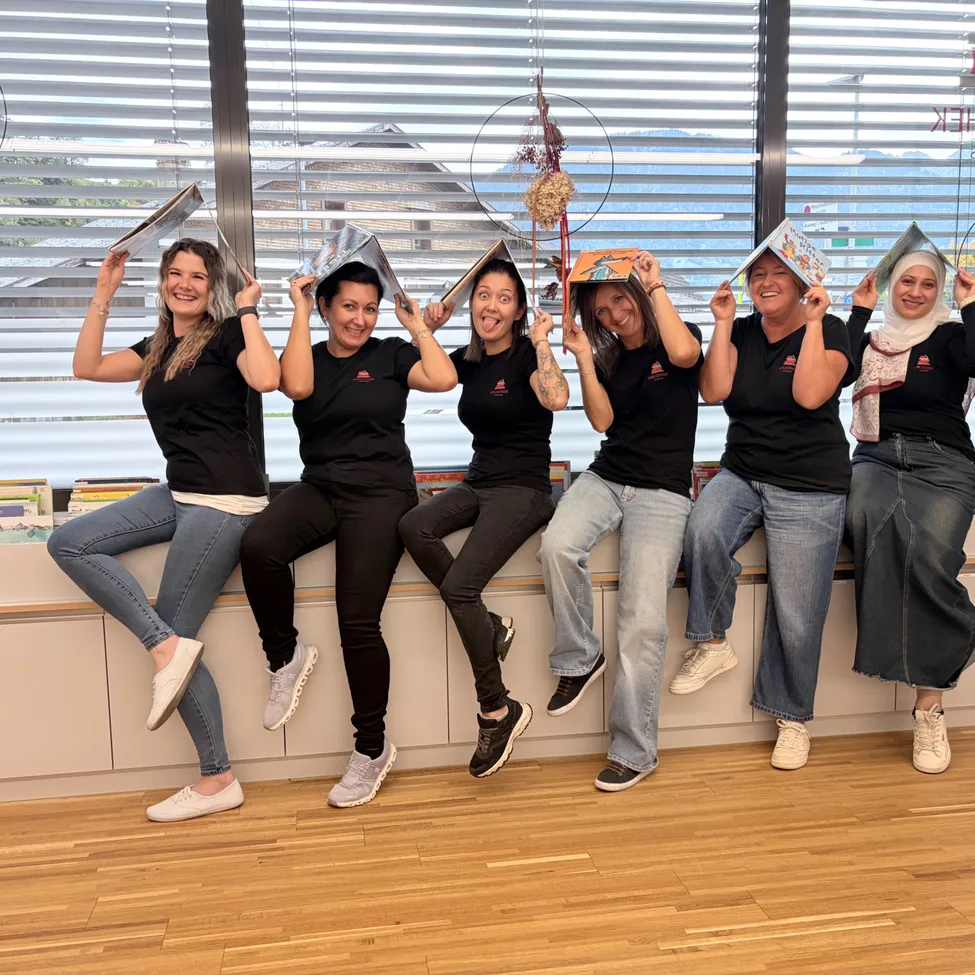Eine Gruppe von Frauen sitzt auf einer Bank und posiert für ein Foto, während sie Bücher über ihren Köpfen halten. Sie tragen alle schwarze T-Shirts und Jeans.