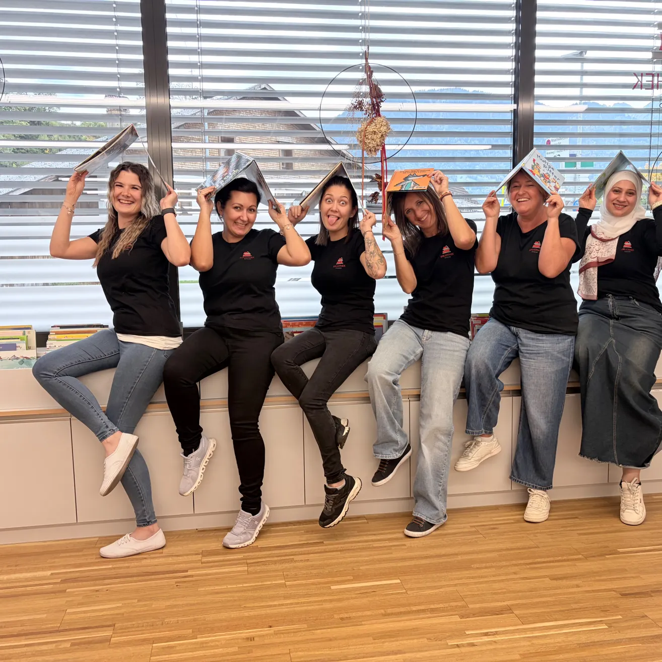 Eine Gruppe von Frauen sitzt auf einer Bank und posiert für ein Foto, während sie Bücher über ihren Köpfen halten. Sie tragen alle schwarze T-Shirts und Jeans.