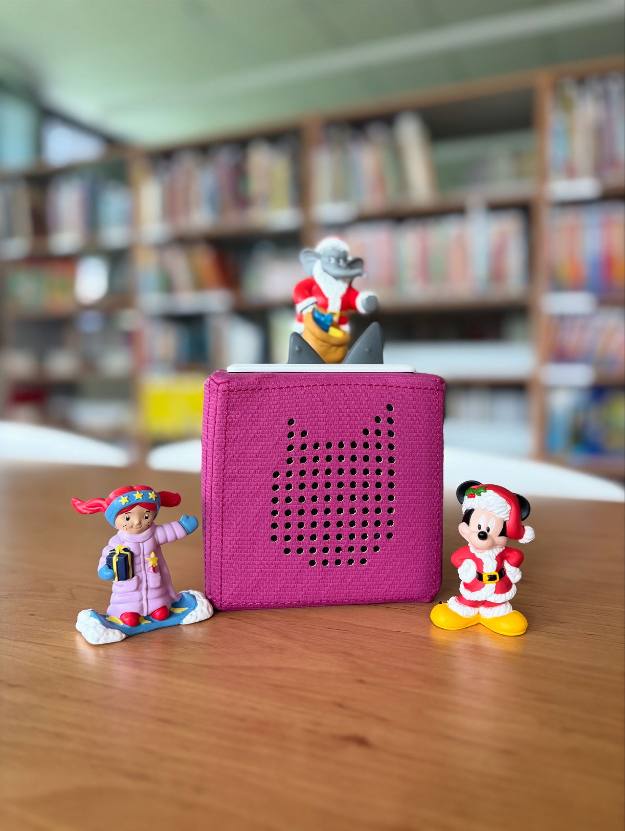 Ein rosafarbener Lautsprecher mit einer Santa-Figur und zwei Spielzeugfiguren auf einem Holztisch in einer Bibliothek.