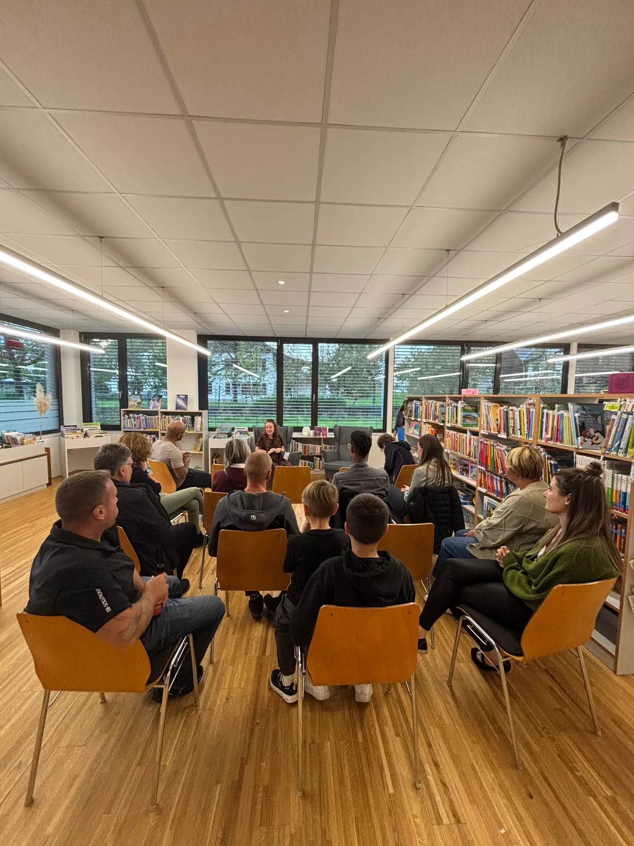 Eine Gruppe von Menschen sitzt in Stühlen um eine Frau in einer Bibliothek und hört ihr zu. Die Bibliothek hat Holzböden, Bücherregale und große Fenster.
