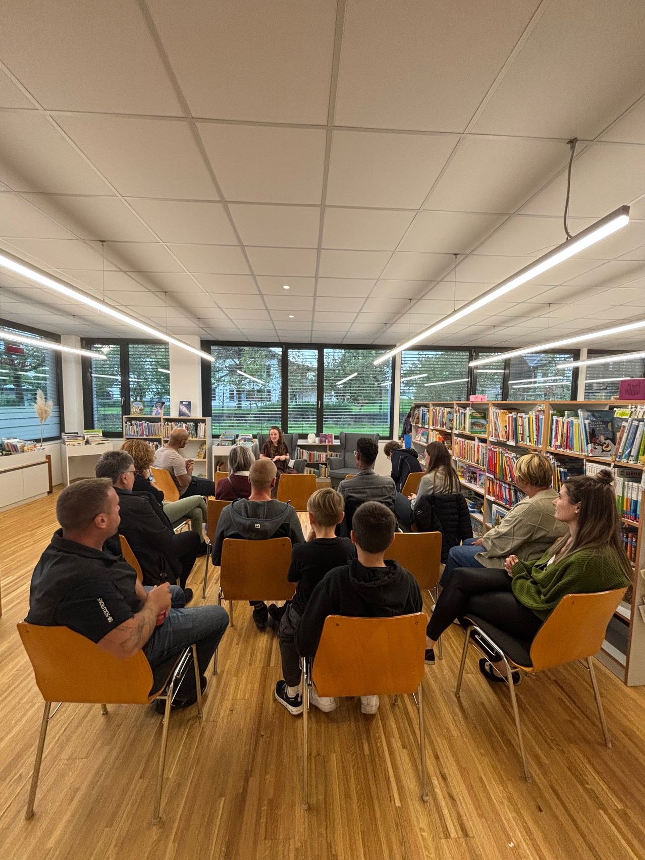 Eine Gruppe von Menschen sitzt in Stühlen um eine Frau in einer Bibliothek und hört ihr zu. Die Bibliothek hat Holzböden, Bücherregale und große Fenster.