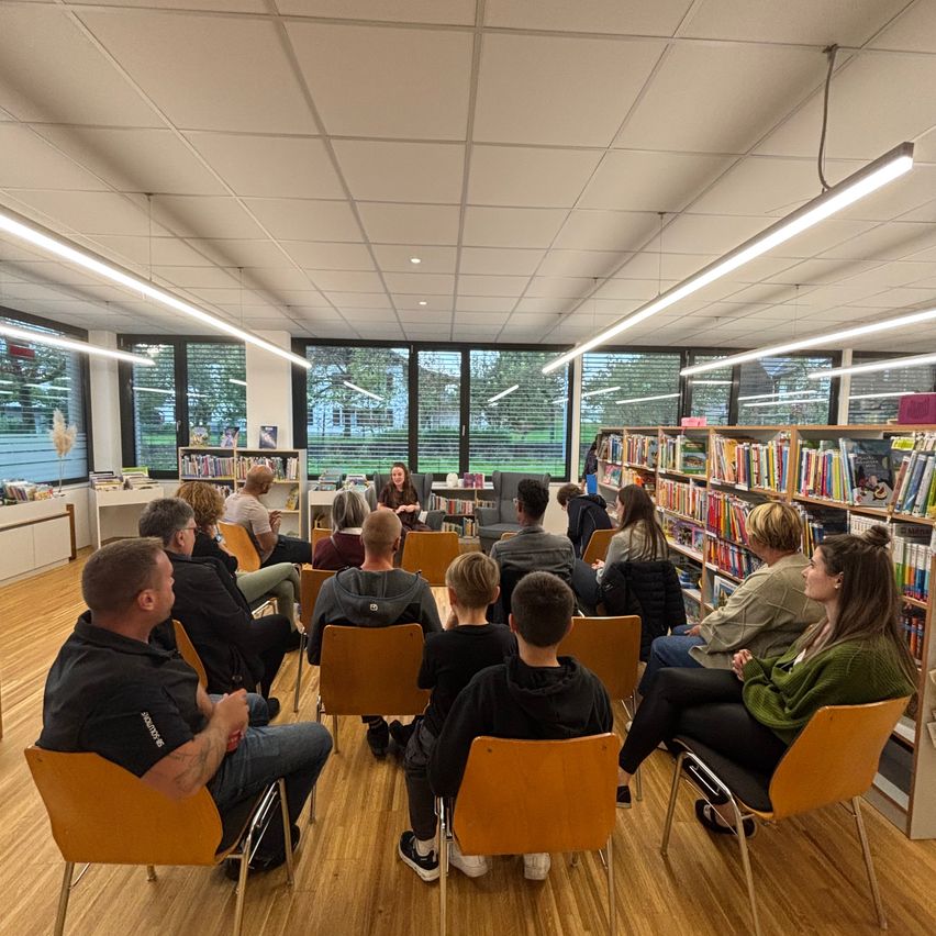 Eine Gruppe von Menschen sitzt in Stühlen um eine Frau in einer Bibliothek und hört ihr zu. Die Bibliothek hat Holzböden, Bücherregale und große Fenster.