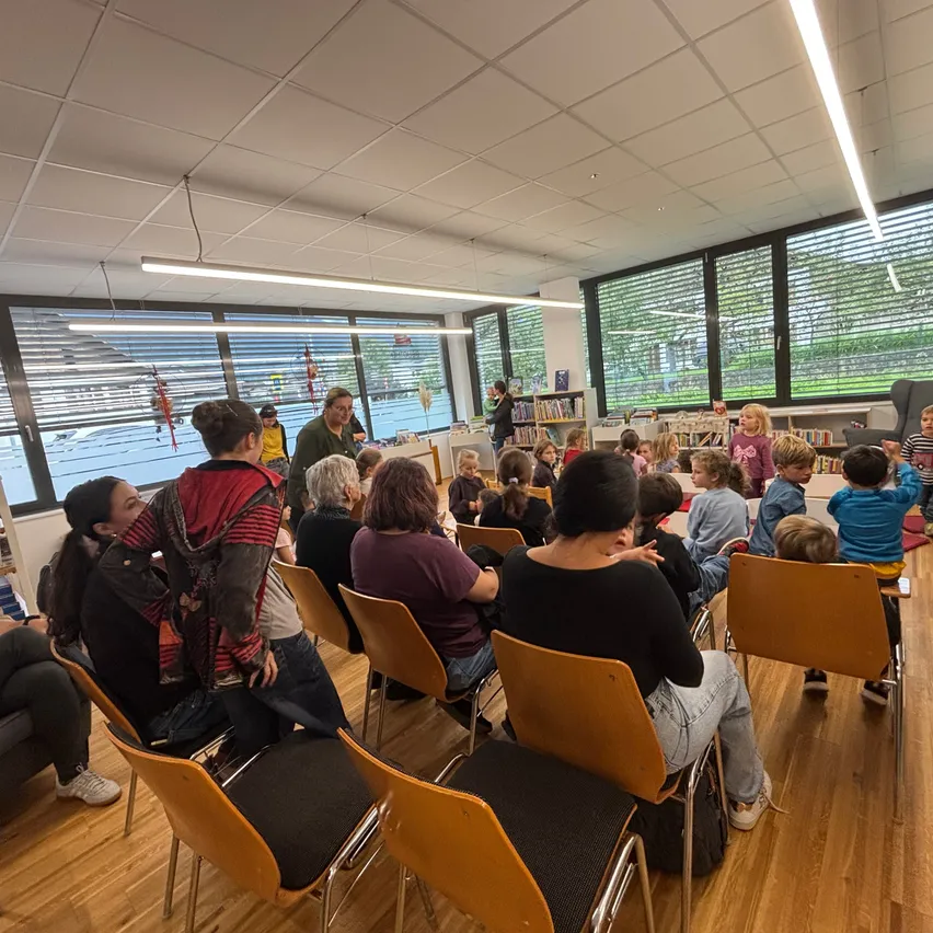 Ein Raum mit einer Gruppe von Menschen, vermutlich Kindern, die auf Stühlen sitzen. Eine Person steht vorne, vermutlich führt sie eine Diskussion. Der Raum hat Glasfenster, Deckenleuchten und einen Teppich auf dem Boden.