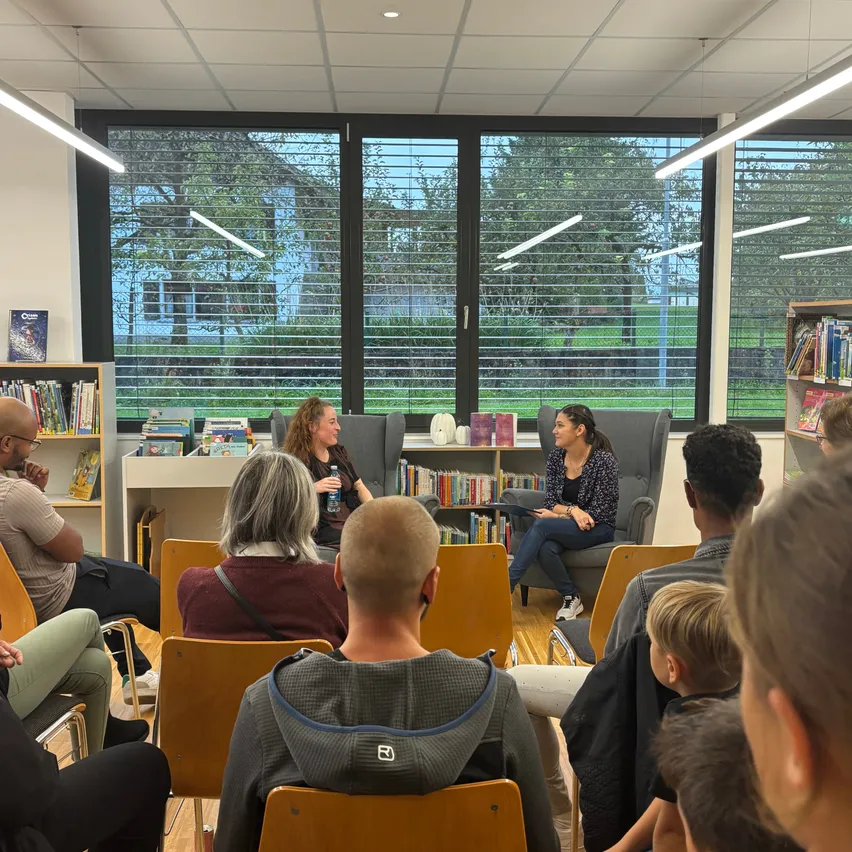 In einer Bibliothek sitzen mehrere Personen in Stühlen um zwei Frauen, eine hält ein Buch. Hinter ihnen sind mit Büchern gefüllte Regale zu sehen. Draußen scheint die Sonne durch die Fenster.