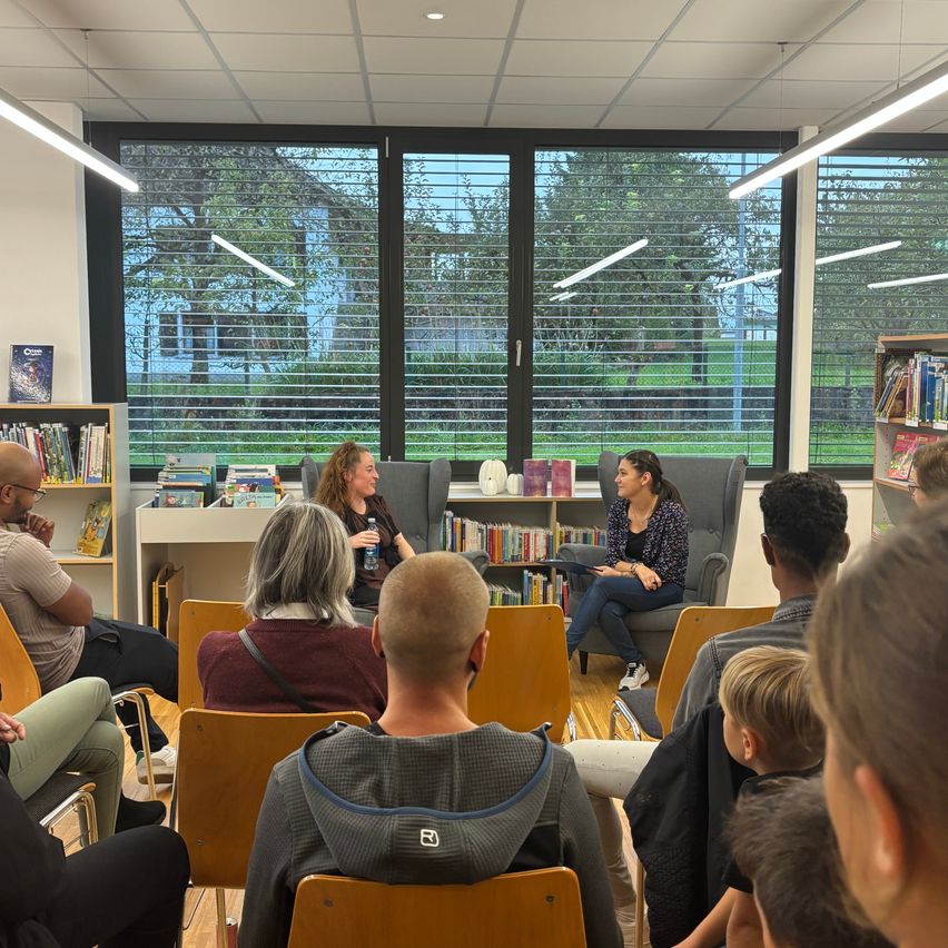 In einer Bibliothek sitzen mehrere Personen in Stühlen um zwei Frauen, eine hält ein Buch. Hinter ihnen sind mit Büchern gefüllte Regale zu sehen. Draußen scheint die Sonne durch die Fenster.