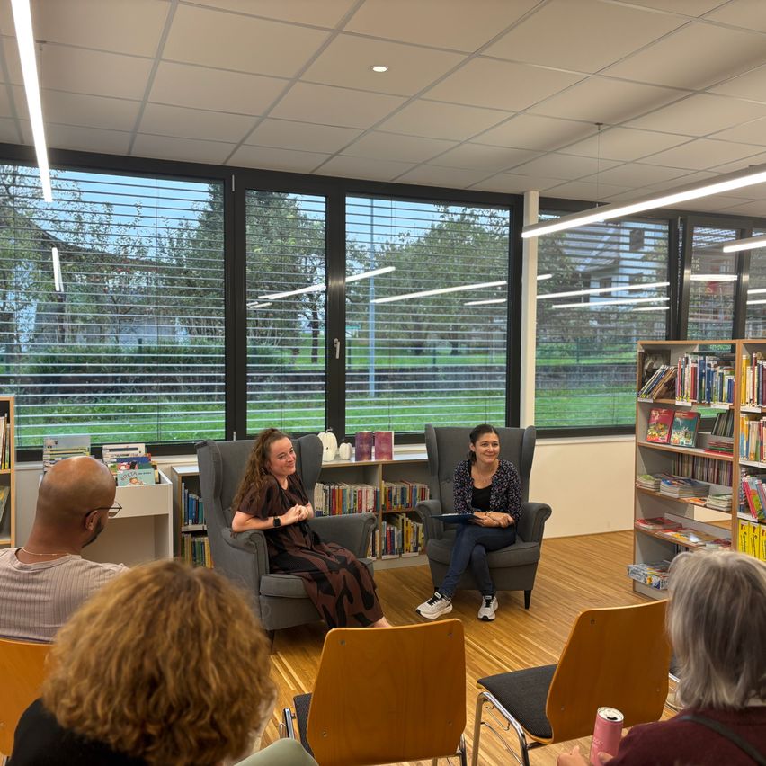 Zwei Frauen sitzen in Stühlen und führen wahrscheinlich ein Interview. Sie sind von einer Gruppe von Menschen umgeben. Der Raum hat Bücherregale, Stühle und Fenster. Außerhalb der Fenster gibt es ein Grasfeld und Bäume.
