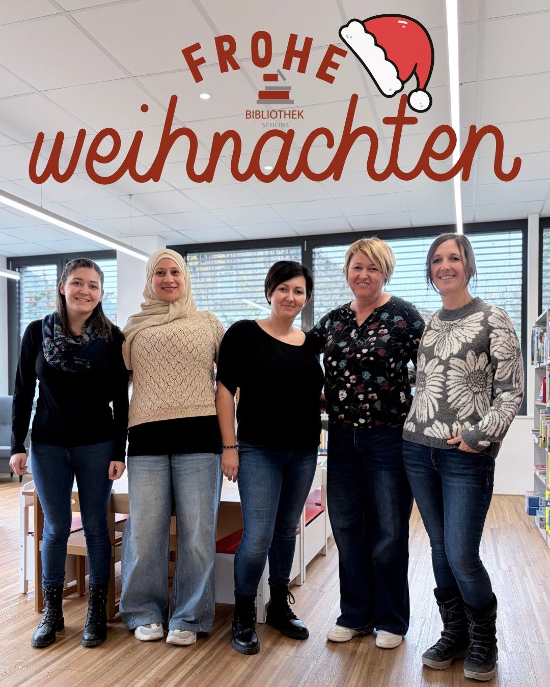 Fünf Frauen posieren lächelnd für ein Foto in einer Bibliothek. Hinter ihnen steht ein festlicher Banner mit der Aufschrift 'Weihnachten' auf Deutsch. Sie stehen vor Bücherregalen und einem Fenster.