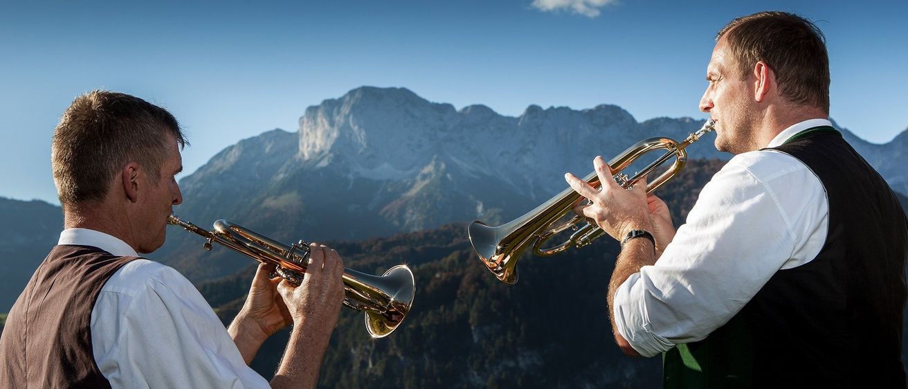 Zwei Männer spielen Trompeten in einem Gebirgsgebiet unter einem blauen Himmel. Die Berge sind mit Bäumen bedeckt.