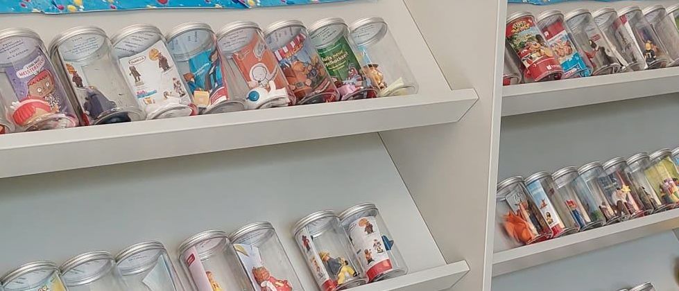 Ein weißer Regal mit durchsichtigen Plastikgläsern, die bunte Spielzeuge und Bücher enthalten, mit einem blauen Himmelshintergrund und einem roten Stuhl auf der rechten Seite.