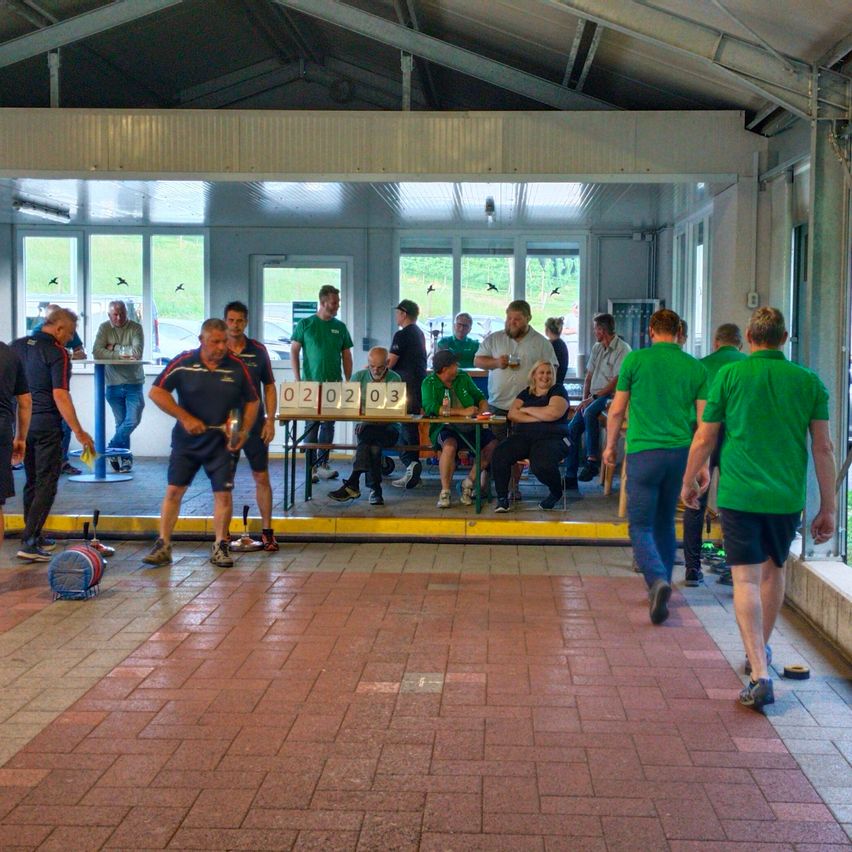 Eine Gruppe von Menschen in grünen Shirts nimmt an einer Sportveranstaltung teil. Einige Leute sitzen an einem Tisch, andere stehen. Ein Mann in der Mitte hält eine Hantel.