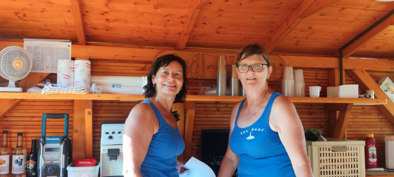 Zwei Frauen in blauen Tanktops stehen in einer Holzhütte, eine lächelt und hält ein Papier in der Hand, die andere trägt eine Brille. Gestackte Becher und ein Holzschild schmücken die Holzwände.