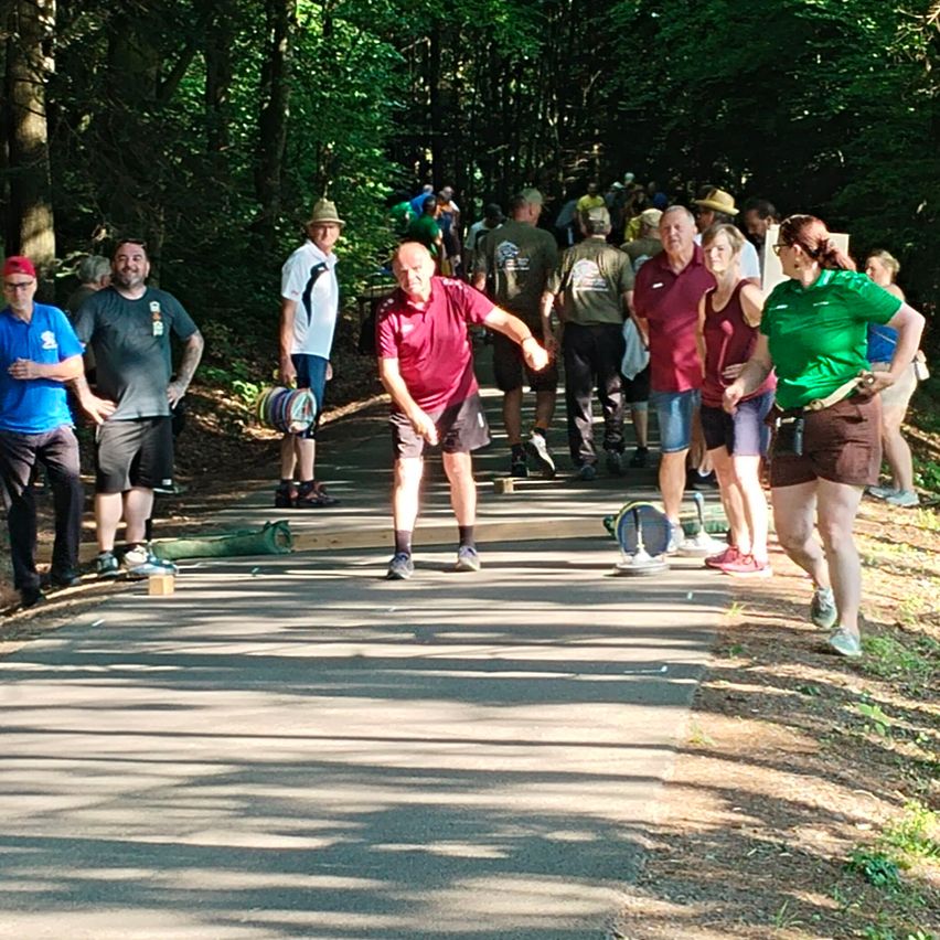 Eine Gruppe von Menschen nimmt an einem Wurfspiel auf einem Betonweg in einem bewaldeten Gebiet teil, wobei einige Spieler in Aktion sind, während andere zusehen.