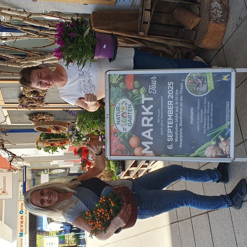 Zwei Frauen posieren für ein Foto, eine hält einen Blumenstrauß in der Hand, und stehen vor einem Schild, das eine Markttour am 6. September 2025 ankündigt.